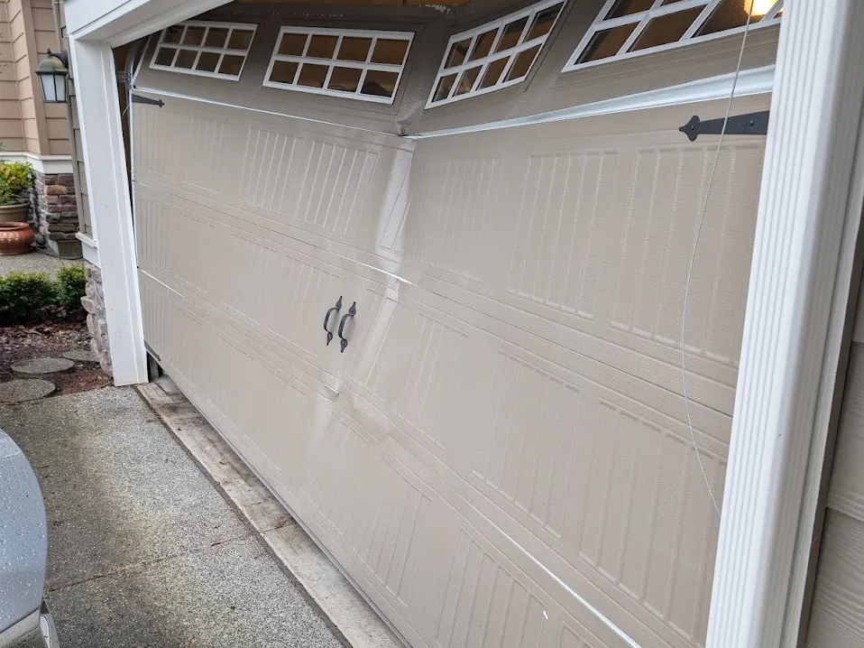 A beige garage door with a buckled panel, sitting crooked in the opening.