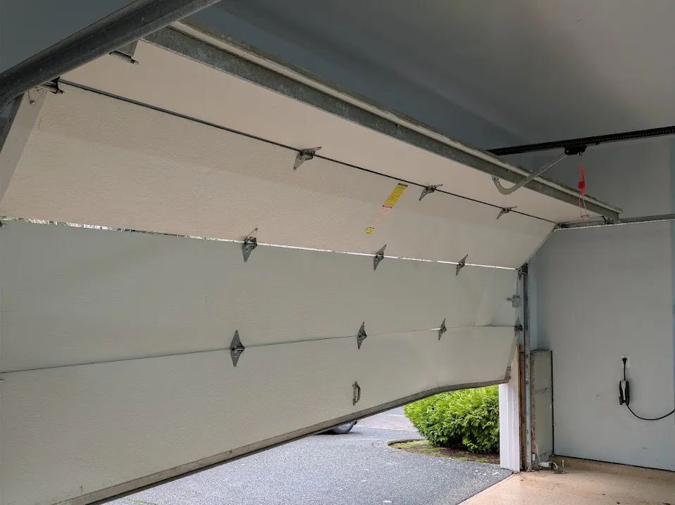 A residential garage door off its track, bowed inward and pulling away from the opening.